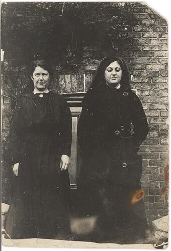 Two woman in dark dresses standing in front of brick building.