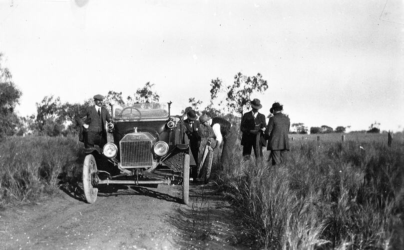 Man mending a tyre on a T Model Ford. Four other men watch.