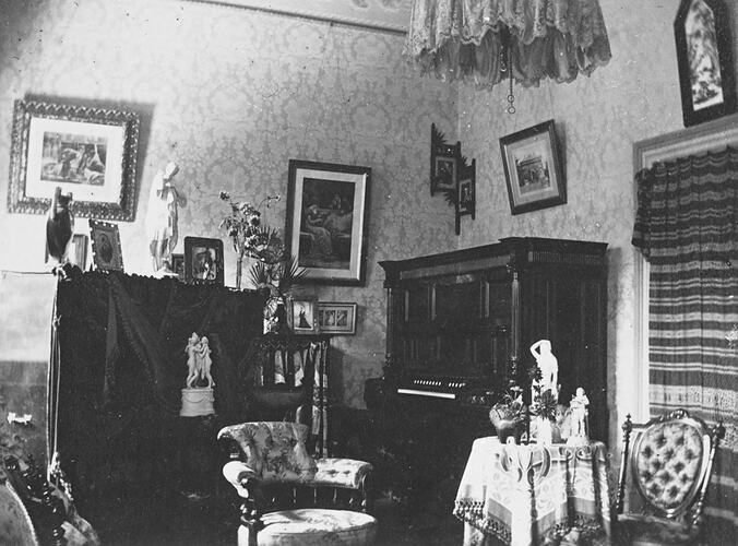 Sitting room with organ at right, pictures on walls, statuettes on tables. Decorative ceiling light.