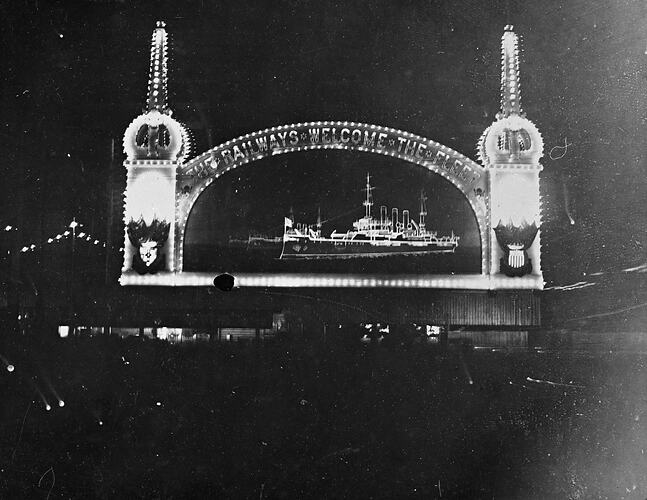 Iluminated arch framing illuminated battleship. The arch carries the legend 'The railways welcomes the fleet'.