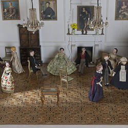 Miniature withdrawing room in a doll's house. Furniture and people inside. Paintings on walls.