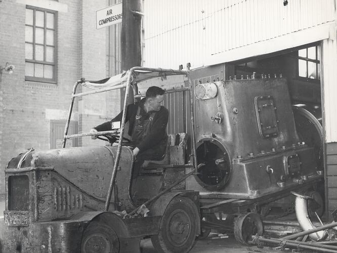 A man is is sitting on a tractor looking at a large compressor which is loaded onto a small trailer behind.