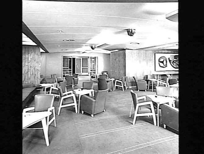 Ship interior. Room with upholstered chairs around tables and some armchairs set up to be a cafe area. Artwork