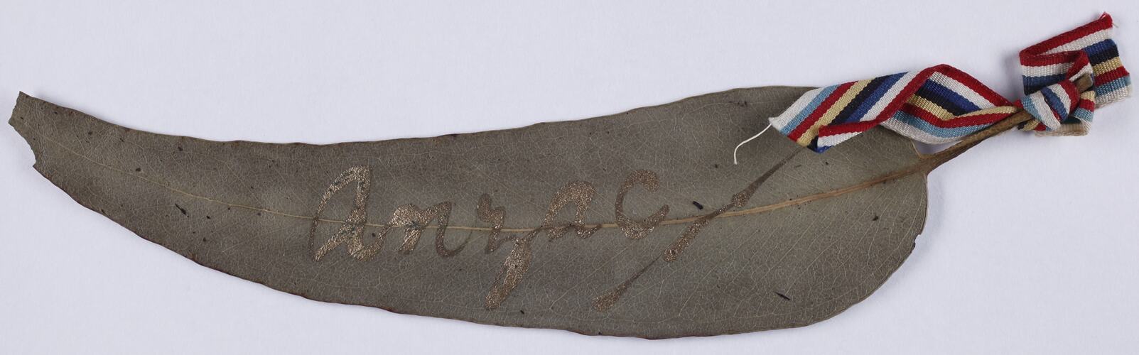 Leaf with handwritten gold text, red, with white and blue ribbon attached to top of leaf,