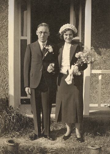 Man and woman wear formal attire in front of house. Woman holds flowers and man has flowers on jacket label.