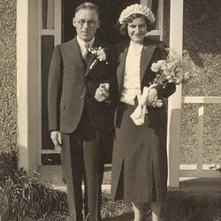 Man and woman wear formal attire in front of house. Woman holds flowers and man has flowers on jacket label.