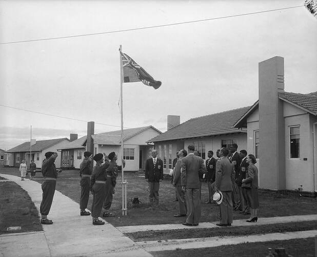 Sportsmen wearing blazers standing to attention around a raised Colonial Flag of Jamaica. Residential buildings in background.