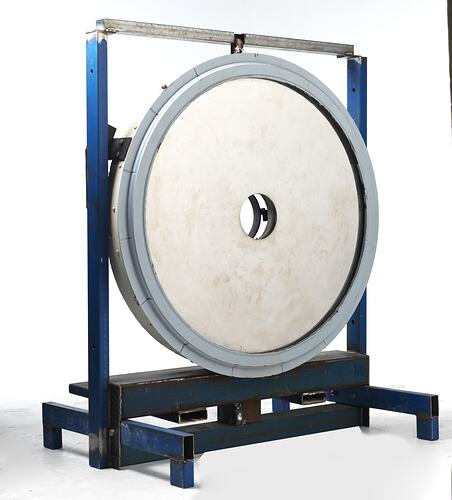 Off-white disc with hole in centre. Grey metal frame. Housed in blue metal rack.