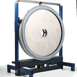 Off-white disc with hole in centre. Grey metal frame. Housed in blue metal rack.
