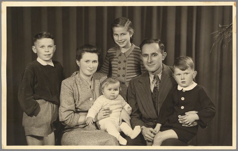 Family portrait of six. Two children stand as two adults sit, the woman holds a baby, the man a young boy.