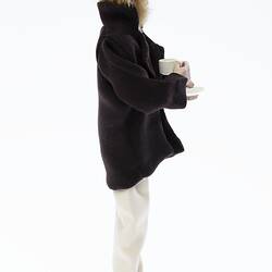 Miniature man in dark coat, light pants. Holds a cup and saucer. Has a hat and fair hair. Profile.