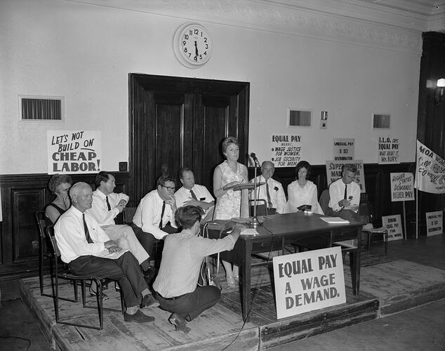 Woman standing at lectern with seated group either side. Poster leans against lectern. Posters on wall behind.