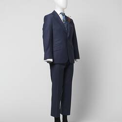 Blue suit jacket and pants with thin red square check pattern. Three outer pockets. Branded brown plastic buttons. White shirt, blue tie. Brown feather corsage. Three quarter view.