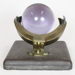 Purple glass sphere sitting on metal frame with square base, rear view.