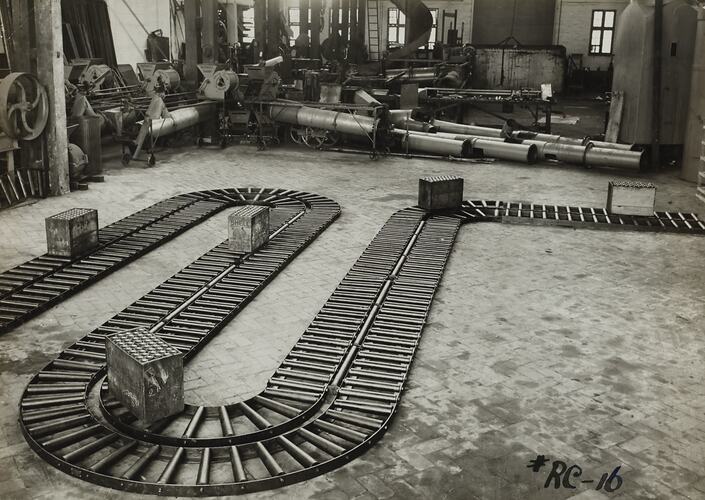 Roller conveyor on floor with crates on top. Pipes stacked behind in factory setting.