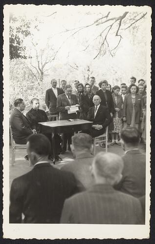 Men sitting at table in front of crowd. Standing group behind them.
