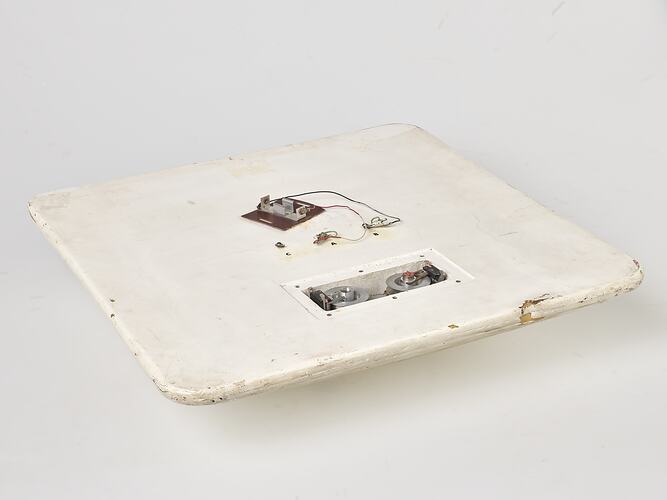 Scuffed white plastic square plate with curved base and rounded corners. Small opening on face side up has two round metal discs within. Above it some wires and a small metal plate are attached to surface.