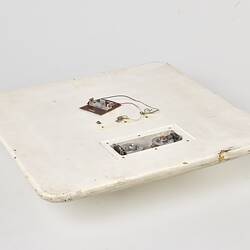 Scuffed white plastic square plate with curved base and rounded corners. Small opening on face side up has two round metal discs within. Above it some wires and a small metal plate are attached to surface.