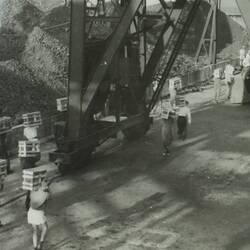 Sailors carrying wooden crates with large steel framed structure on road.