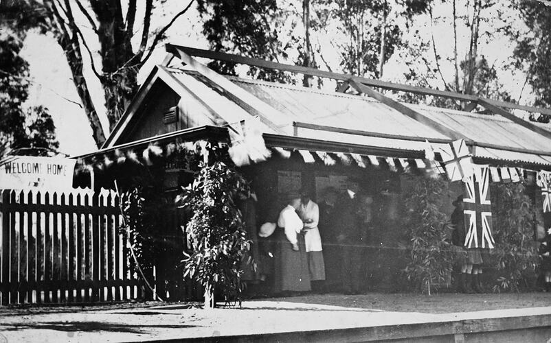 Small crowd standing on railway station decorated with flags and bunting. Features a 'welcome home' banner.