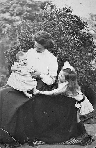 Woman sitting outside with a young child on her lap and a small girl by her side.