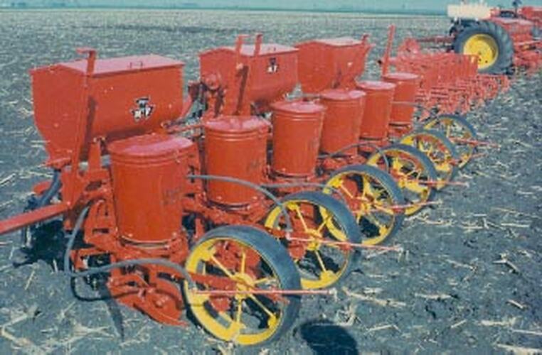 Side view of 6 seed planters and 3 fertilizer bins on a tool bar with ploughed field in background.