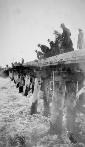 People standing on a narrow bridge looking down at the floodwaters below.