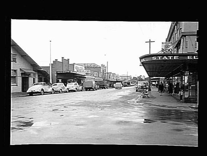 Street view with cars parked on sides of road in front of shops and businesses.