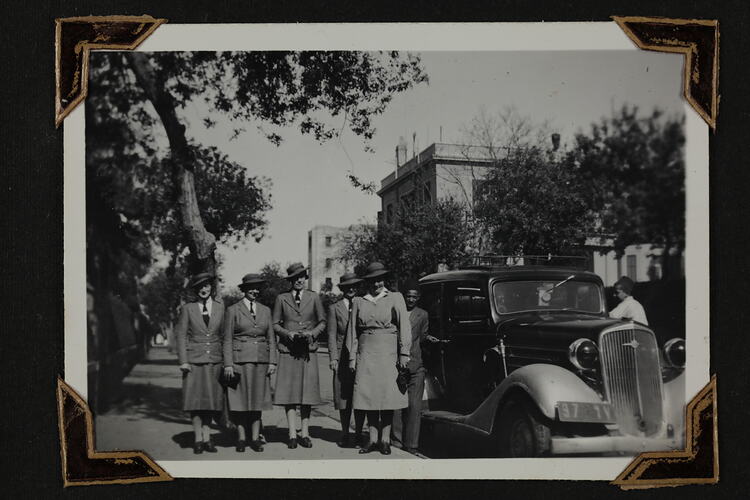 Group of five uniformed woman and two men standing beside motor vehicle on street.