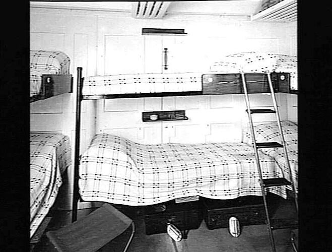Ship interior. Bunk beds with checked blankets. Ladder at right.