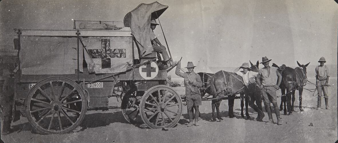 Horse-drawn carriage with red cross on side, four men on standing ground by horses.