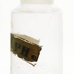 Glass jar with glass stopper. Painted label in metallic and black inks. Contains small white flakes.