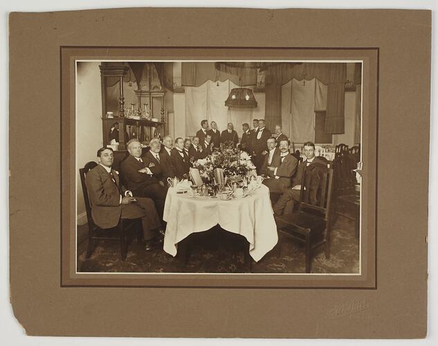 Group of seated and standing men at formal dinner setting.