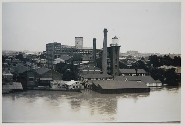 View from a riverbank looking across the flooded water to partially submerged factories and buildings. Two chi