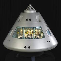Conical shaped silver spacecraft model. Three seated astronauts inside facing upwards.