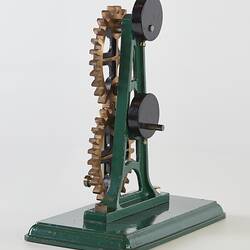 Model of two brass coloured metal elliptical gear wheels on a frame with a handle, mounted on green base.