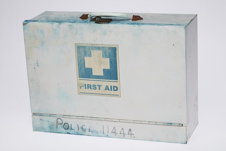 White metal case with handle on top. Side has green square with white cross. Text below.