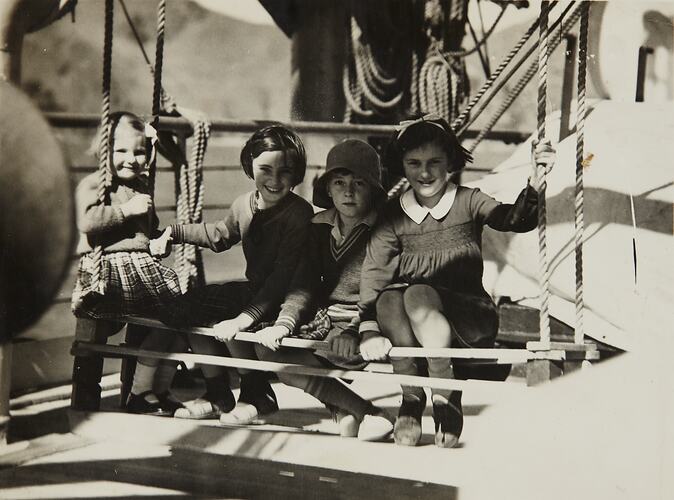 Four children sit in a row on wooden planks suspended by ropes.