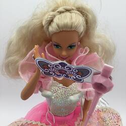 Detail of Barbie doll with layered pink and white dress. She holds a fancy dress mask.