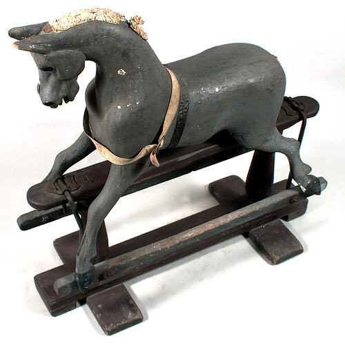 Dark grey wooden rocking horse with pale cloth or woollen mane. Brown belt around neck. Base is dark brown.