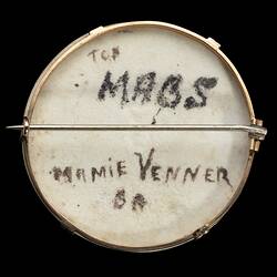 Back of white porcelain brooch with gold frame and horizontal pin. Black handwriting in centre.