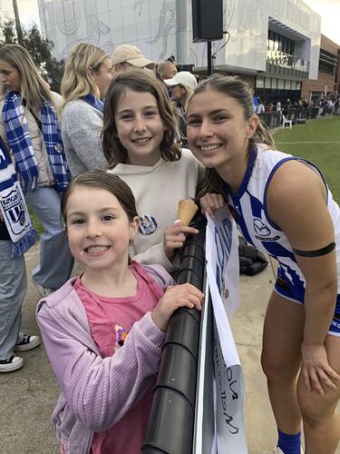 Two girls posing with female footballer. They stand on left side of low fence, footballer stands on right side. They are smiling at camera.
