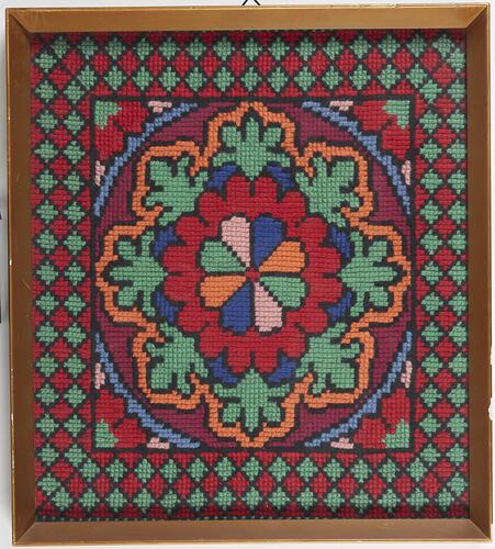 Framed square cloth embroidered with central 8-petalled floral motif in blue, green, yellow and light pink. Red, green and yellow patterns form border.