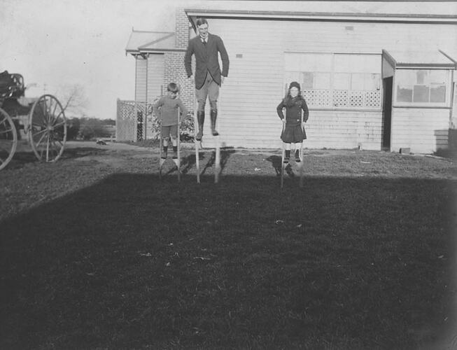 Digital Photograph - Man, Boy & Girl Walking on Stilts, Farmhouse Yard, Won Wron, circa 1915