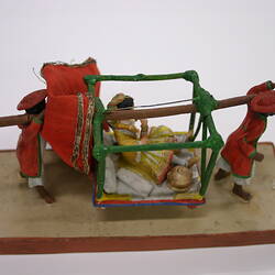 Small model of two men carrying a pole bearing a hanging seat with a lady reclined on cushions