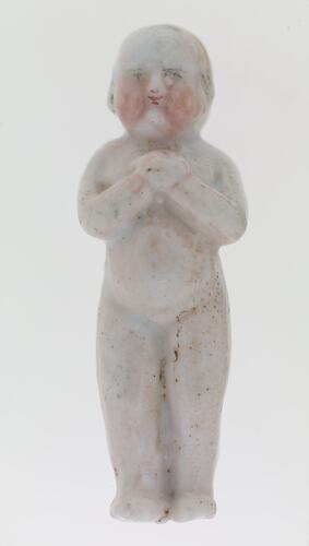 Porcelain doll with hands clasped over its chest. Painted eyes, nose, mouth and hair are faded and worn.