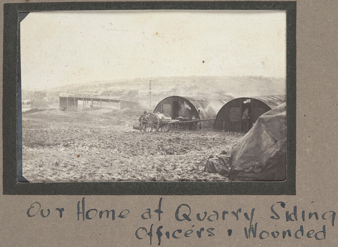 Two wooden huts with cart in front, serviceman in entrance to one hut, structures visible in background.