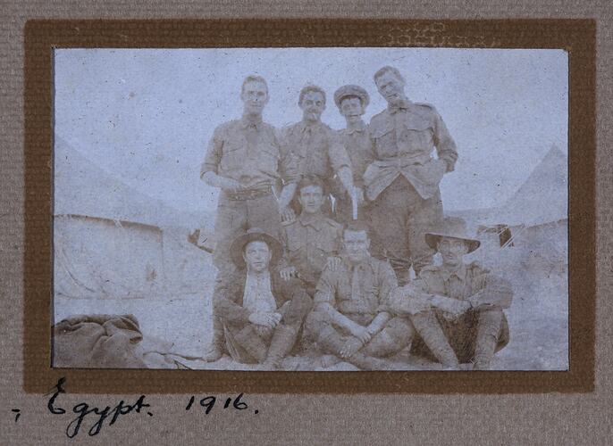 Group of eight servicemen, seated and standing, with tents behind.
