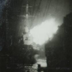Military battleship with explosion in the air to right of ship, ocean in the foreground.