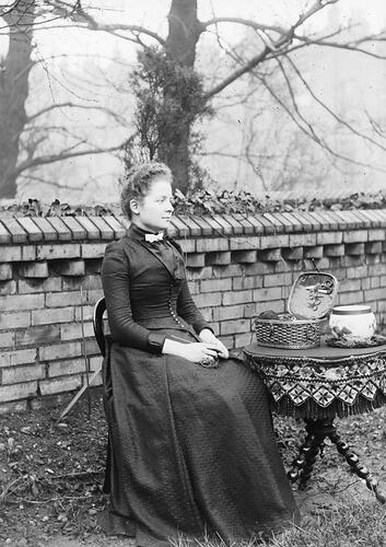 Young woman in black dress sitting in front of a brick wall with a table beside her.